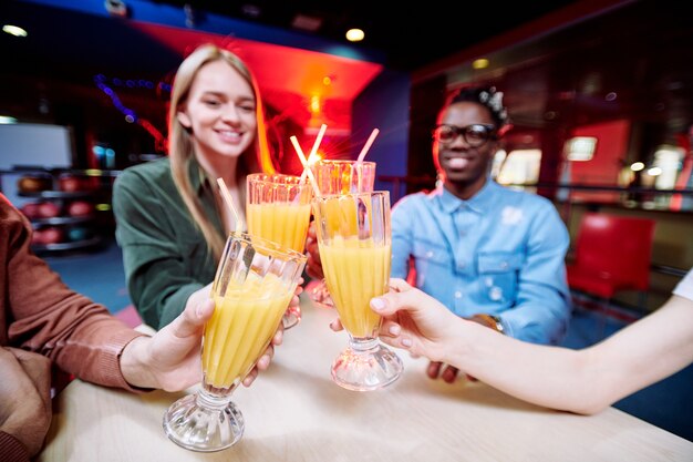 Quatro jovens amigos interculturais tilintando com copos de suco de laranja fresco à mesa enquanto passam um tempo no café do centro de lazer