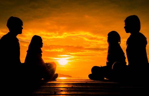 Quatro jovens sentam-se no cais de madeira ao nascer do sol na praia para fazer meditação com laranja lindo céu e nuvens.