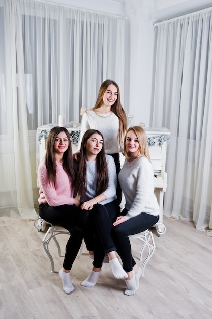 Quatro meninas amigos bonitos usam blusas quentes e calças pretas contra piano velho com decoração de Natal na sala branca.