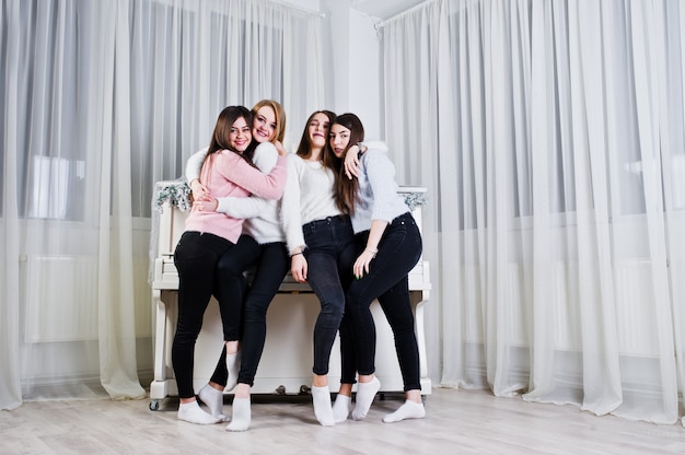 Quatro meninas amigos bonitos usam blusas quentes e calças pretas contra piano velho com decoração de Natal na sala branca.