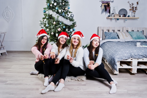 Quatro meninas amigos bonitos usam camisolas quentes, calças pretas e chapéus vermelhos de papai noel contra a árvore com decoração de natal no quarto branco