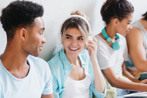 Foto que bom que menina com cabelo loiro tocando seus fones de ouvido brancos e olhando com sorriso para um jovem africano bonito. aluna bonita curtindo música e passando tempo com os amigos.