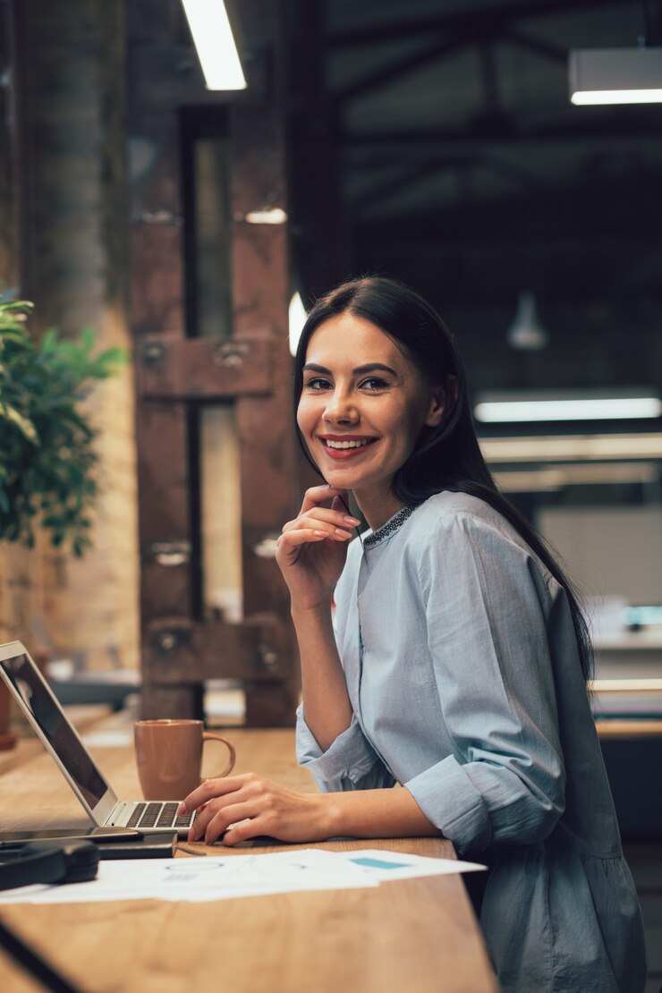 Que bom que moça bonita tocando seu queixo e sorrindo enquanto está sentada com um laptop na mesa