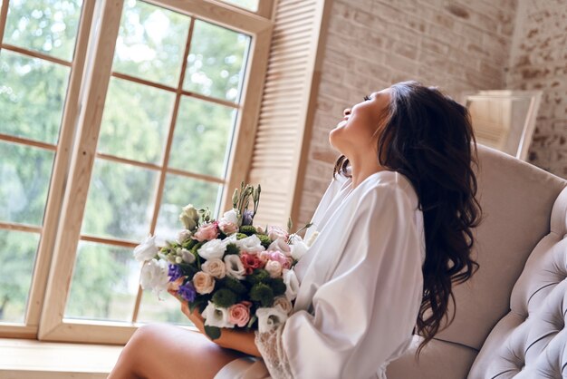 ¡Qué bonita mañana! Hermosa mujer joven en bata de baño de seda sosteniendo un ramo y sonriendo mientras está sentado en el sofá