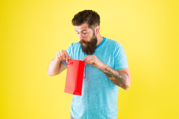 Qué bonita tienda sorpresa para hombres Barba masculina madura con compra Hipster sorprendido Sostenga la bolsa de papel roja Mejor oferta Hombre con paquete Lunes cibernético Poca amabilidad Hombre barbudo Ir de compras