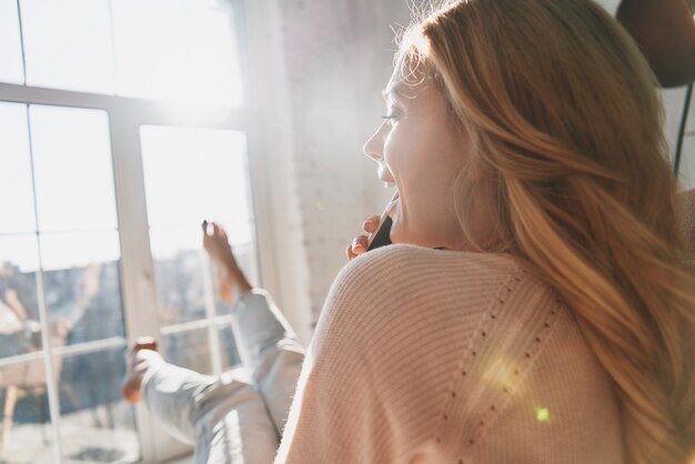 ¡Qué bueno escucharte! Vista trasera de la hermosa joven hablando por su teléfono inteligente y sonriendo mientras está sentado cerca de la ventana en casa