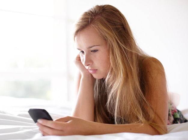 ¿Qué debo decirle? Foto de una hermosa joven leyendo un mensaje de texto mientras está acostada en su cama.