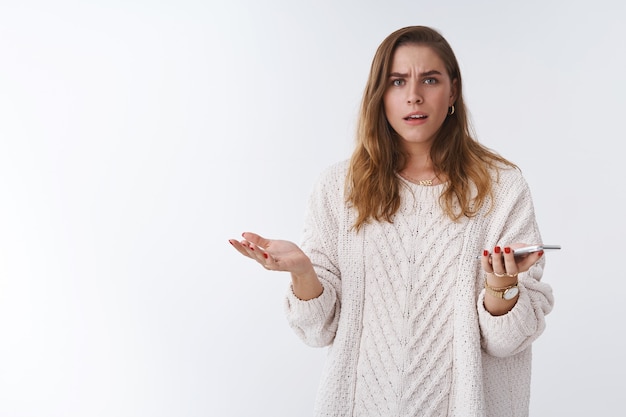 ¡Qué diablos! Retrato frustrado enojado cuestionado mujer despistada discutiendo sosteniendo teléfono inteligente encogiéndose de hombros con las manos extendidas consternación frunciendo el ceño perplejo boca abierta hablando intenso, fondo blanco