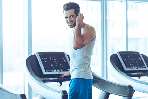 ¿Qué dijiste? Vista trasera del joven apuesto en ropa deportiva de pie en la caminadora frente a la ventana en el gimnasio y ajustando sus auriculares mientras mira a la cámara con una sonrisa