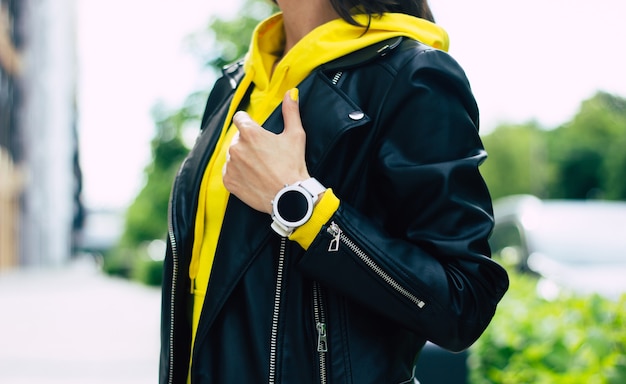 ¡Qué diseño tan agradable! look de mujer joven, que consta de chaqueta de cuero, sudadera con capucha amarilla y su nuevo reloj inteligente bien diseñado.