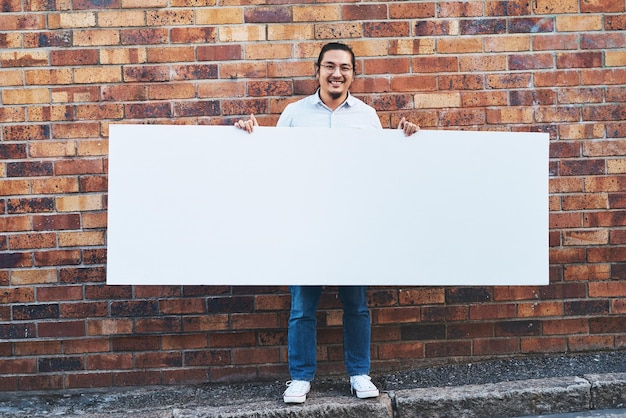 ¿Qué harías con todo este espacio? Foto de un joven sosteniendo una pancarta en blanco contra un fondo urbano al aire libre.