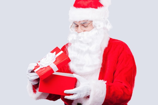 ¿Qué hay adentro? Papá Noel tradicional sosteniendo una caja de regalo y mirando dentro de ella mientras está de pie contra el fondo gris