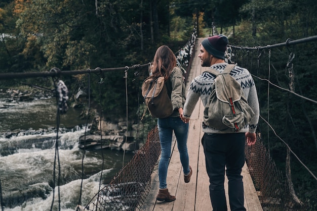 ¿Qué hay al otro lado? Vista trasera de la joven pareja cogidos de la mano mientras camina sobre el puente colgante