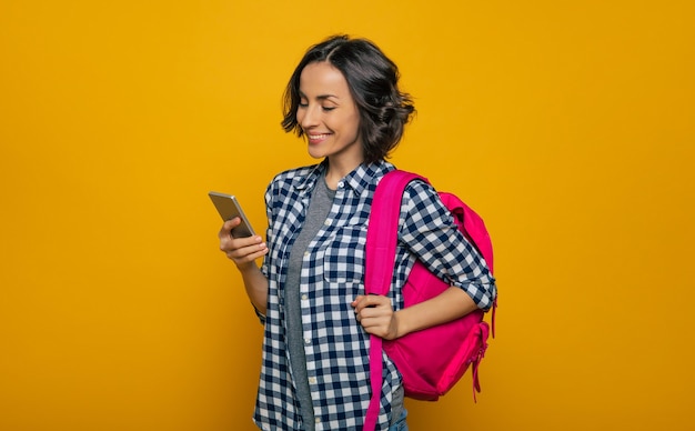 ¿Qué hay de nuevo en Internet? Una hermosa joven estudiante, vestida de manera informal, con su linda mochila rosa al hombro, mirando con una amplia sonrisa en su teléfono inteligente.