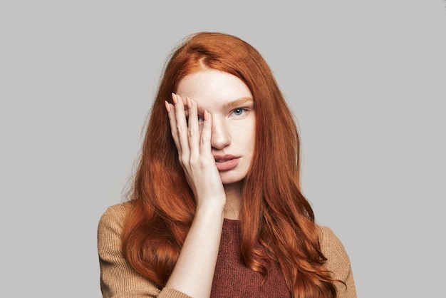 Foto ¿qué hay? retrato de una joven y linda adolescente con cabello rojo sedoso cubriendo la cara con la mano y mirando a la cámara mientras está de pie contra el fondo gris