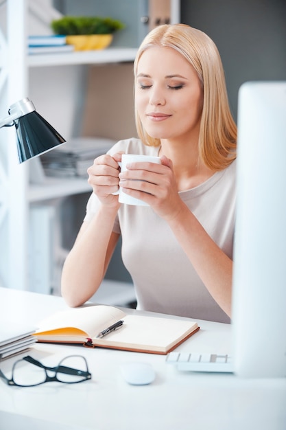 ¡Qué hermoso día! Hermosa joven sosteniendo una taza con bebida caliente y manteniendo los ojos cerrados mientras está sentada en su lugar de trabajo en la oficina