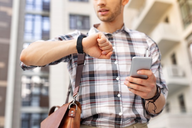 Qué hora es. Apuesto joven mirando la hora mientras sostiene su teléfono inteligente en la mano