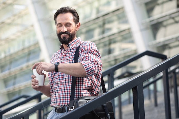 Qué hora es. Hombre barbudo alegre comprobar el tiempo mientras bebe café al aire libre