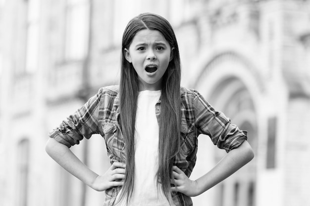 Qué niña frustrada con cabello largo usa estilo casual manteniendo la boca abierta y las manos en las caderas chica de verano al aire libre