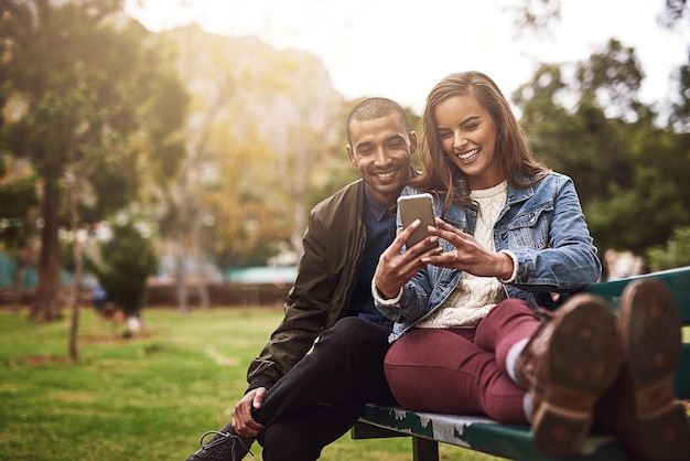 Foto ¿qué opinas de esta foto? la foto de una pareja joven y alegre sentada en un banco mientras usan un teléfono juntos afuera en un parque.