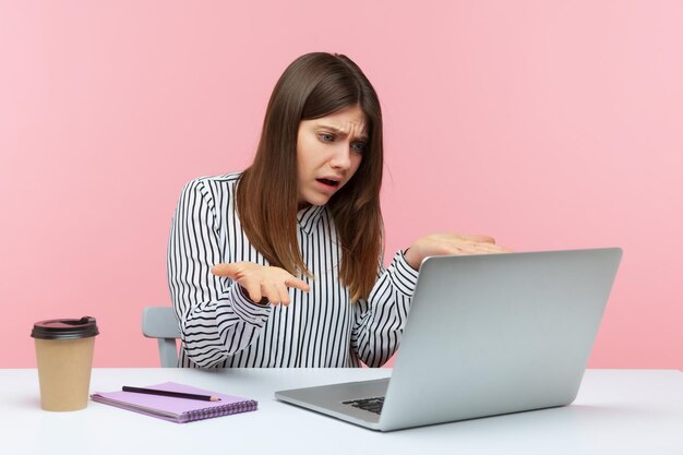 ¿Qué quieres? Una mujer de negocios enojada y nerviosa extendiendo las manos haciendo preguntas mirando la pantalla de un portátil en una conferencia en línea con un compañero en un estudio interior aislado en un fondo rosa