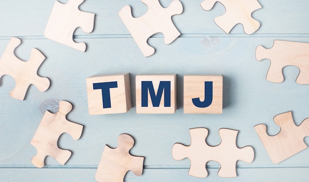 Quebra-cabeças em branco e cubos de madeira com o texto TMJ mentem sobre um fundo azul claro.