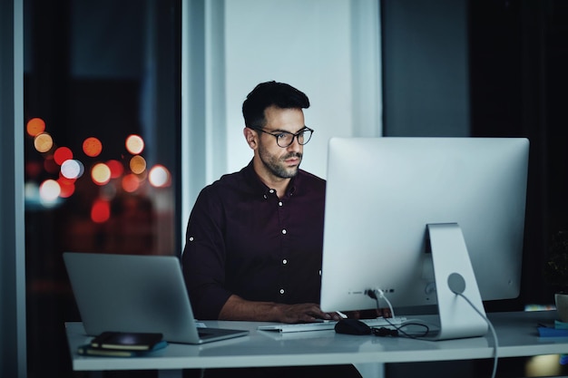 Quedarse hasta tarde para hacerlo Captura de un joven hombre de negocios usando una computadora en su escritorio durante una noche en el trabajo