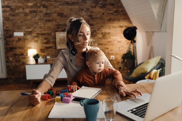 Quédese en casa madre trabajando en una computadora portátil mientras cuida a su hijo pequeño
