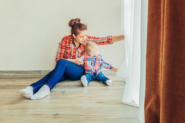 Quédese en casa, manténgase seguro. Joven madre sosteniendo a su hijo. Mujer y niña infantil relajándose en la habitación blanca cerca de windiow en el interior. Familia feliz en casa. Mamá joven jugando con su hija.