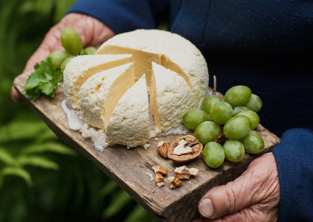 Queijo cottage em uma bandeja de madeira, com uvas, uma noz, nas mãos de uma senhora idosa