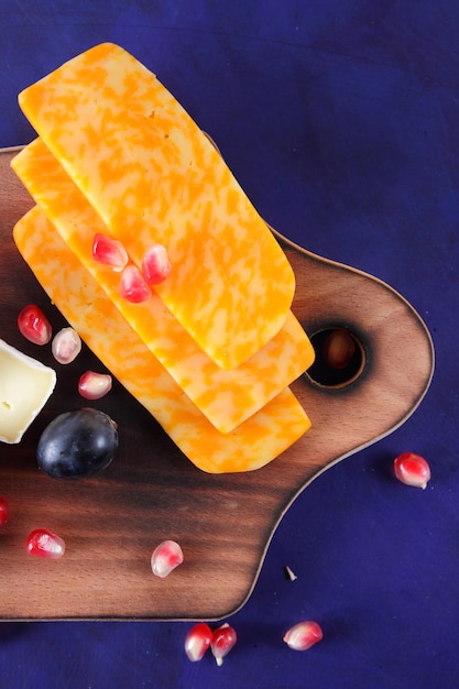 Queijos deliciosos em uma placa de madeira closeup Camembert e fatias de queijo duro amarelo com sementes de romã e uvas escuras em um fundo azul