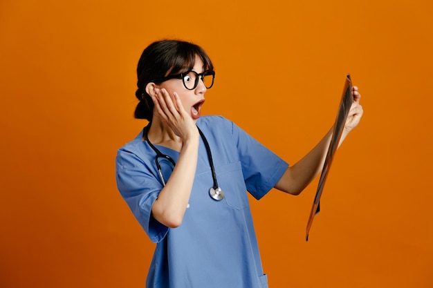 Queixo agarrado insatisfeito segurando a jovem médica de raio-x usando estetoscópio uniforme de quinto isolado em fundo laranja