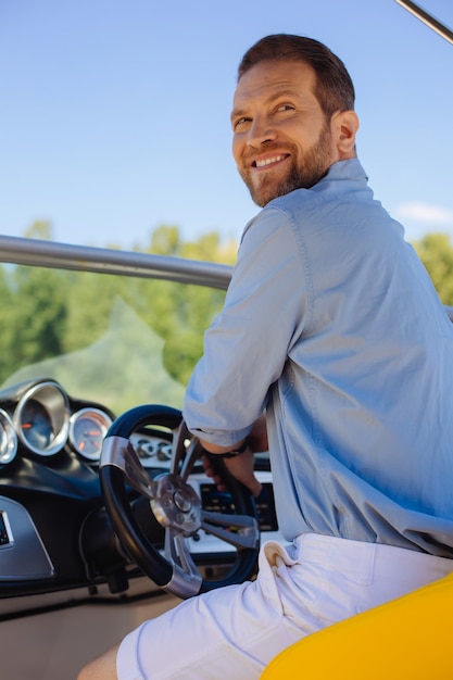 Quem está aqui. Jovem encantador sentado atrás do volante de um iate e se virando com um sorriso nos lábios ao ser chamado por alguém