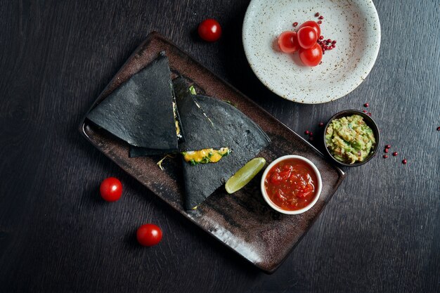 Quesadilla assado apetitoso com 4 tipos de queijo em tortilha preta em um prato de cerâmica em uma mesa preta ... cozinha mexicana moderna.