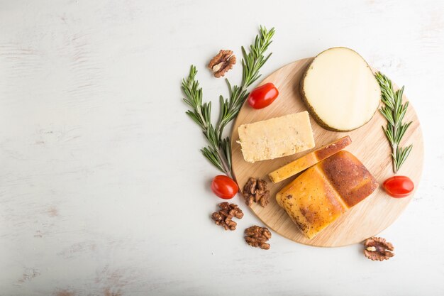 Queso ahumado y diversos tipos de queso con romero y tomates sobre tabla de madera sobre una superficie de madera blanca