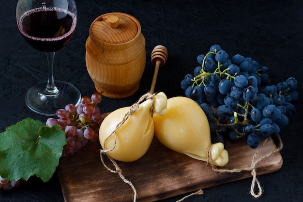 Foto queso caciocavallo en un tablero de madera con las uvas, la miel y el vino en un fondo negro. pera de queso