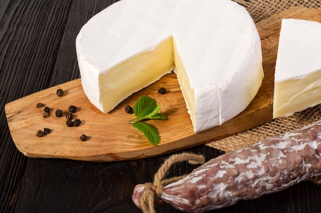 Foto queso camembert redondo, en rodajas, sobre una tabla de madera y salchicha fuet.