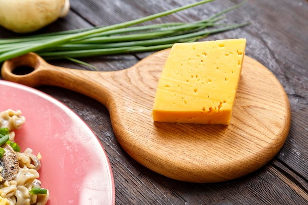 Queso en una tabla de madera junto a cebollas verdes en la mesa y un fragmento de un plato con pasta en salsa cremosa