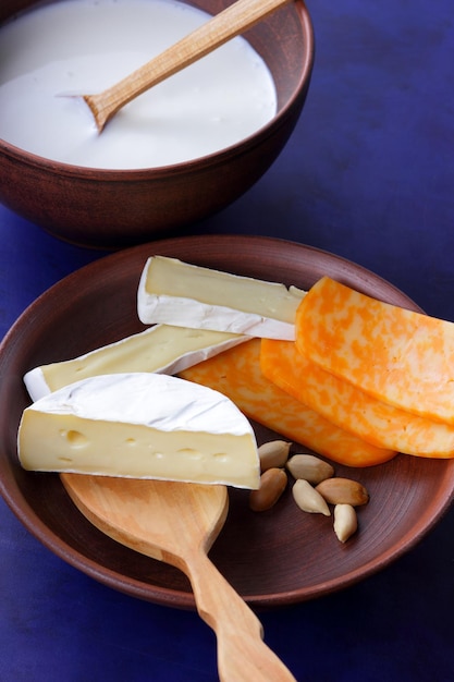 Quesos con nueces y una espátula de madera sobre un plato de arcilla Diferentes tipos de plato de queso y un tazón de leche sobre un fondo azul cerrado