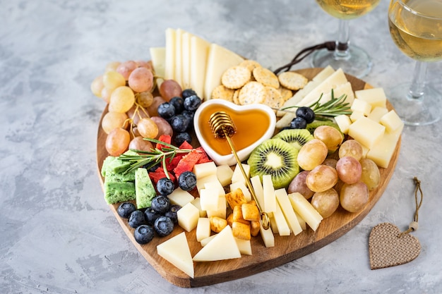 Quesos surtidos sobre una tabla de cortar de madera en forma de corazón. Queso, uvas, nueces, aceitunas, romero y una copa de vino blanco.