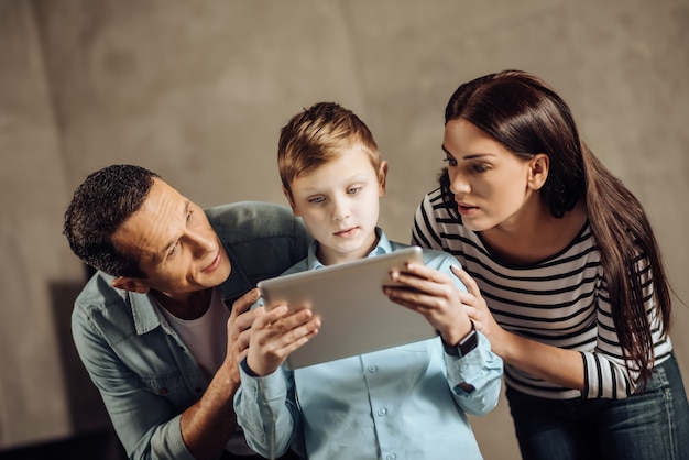 Questão séria. Pais jovens amorosos ao lado de seu filho pré-adolescente e gentilmente o persuadindo a parar de jogar no tablet enquanto o menino não dá atenção a eles