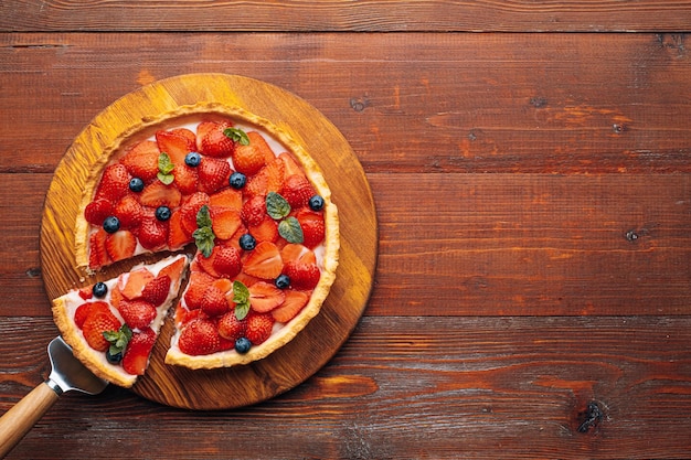 Quiche con ralladura de lima en la que se cortan fresas en rodajas con arándanos y hojas de menta fresca sobre una mesa de madera, se corta una rebanada del pastel y está lista para servir