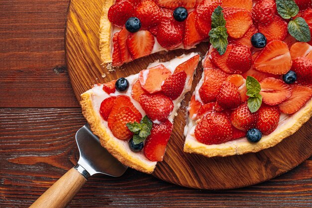 Quiche con ralladura de lima en la que se cortan fresas en rodajas con arándanos y hojas de menta fresca sobre una mesa de madera, se corta una rebanada del pastel y está lista para servir