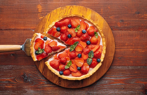 Quiche con ralladura de lima en la que se cortan fresas en rodajas con arándanos y hojas de menta fresca sobre una mesa de madera, se corta una rebanada del pastel y está lista para servir