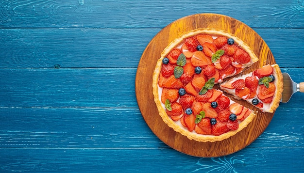 Quiche con ralladura de lima y rodajas de fresa con arándanos y hojas de menta fresca sobre una mesa de madera azul, se corta una rebanada del pastel y está lista para servir