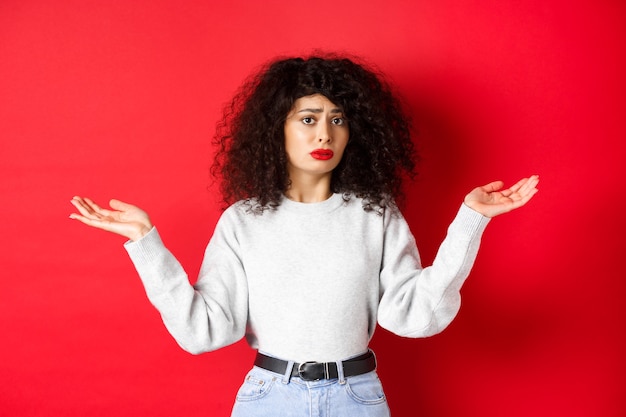 Quién sabe. Mujer joven despistada con el pelo rizado, encogiéndose de hombros y mirando confundido, de pie en ropa casual sobre fondo rojo.