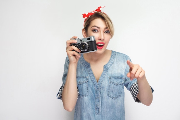 Quieres hacer photoshout. Chica joven blogger en camisa de mezclilla casual con maquillaje, diadema roja de pie, sosteniendo una cámara retro y señalando con el dedo. Foto de estudio de interior, aislado sobre fondo blanco.