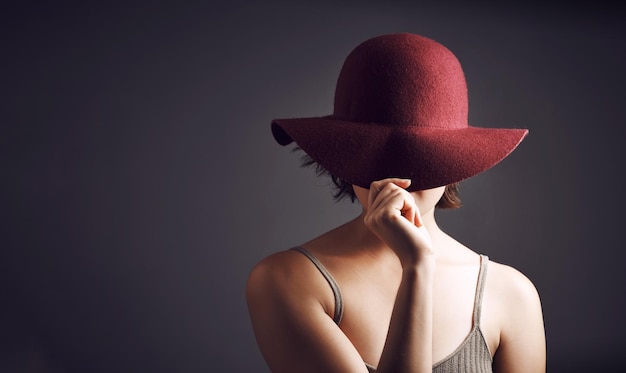 ¿Quieres que revele la misteriosa foto de estudio de una joven irreconocible que se cubre la cara con un sombrero sobre un fondo gris?