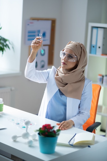 Química feminina. Química feminina usando hijab e uniforme sentada e olhando para o tubo de ensaio