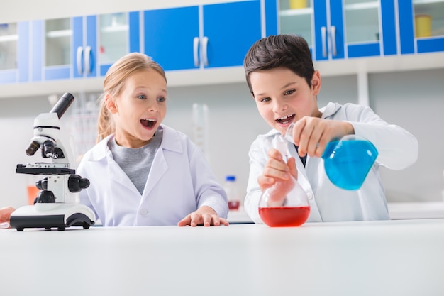 Química interesante. Chico inteligente alegre positivo sosteniendo matraces y mezclando reactivos químicos mientras está en el laboratorio